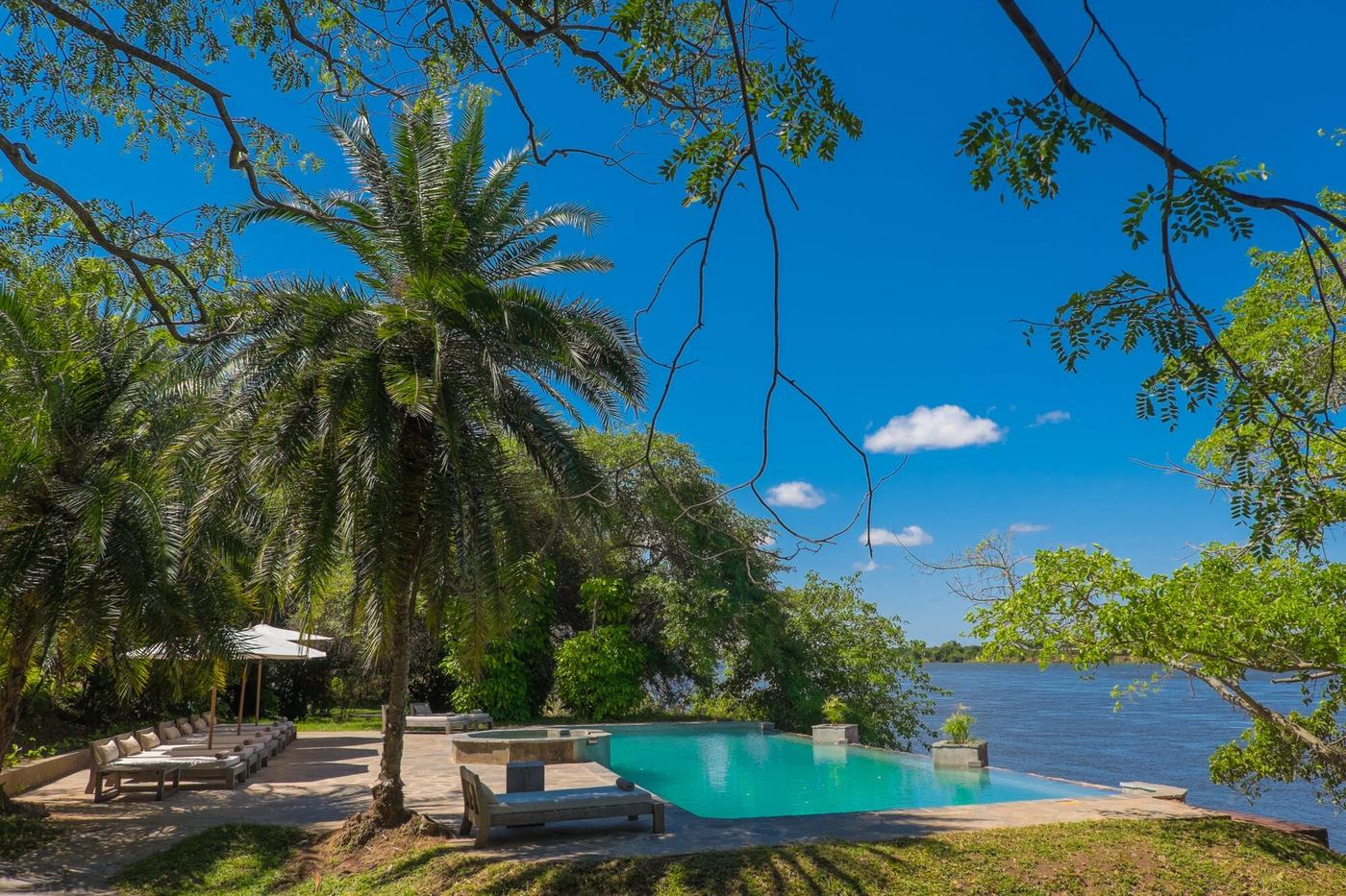Ein Infinity-Pool und Liegen befinden sich unter Palmen mit Blick auf die Zambezi in Kanyemba Lodge