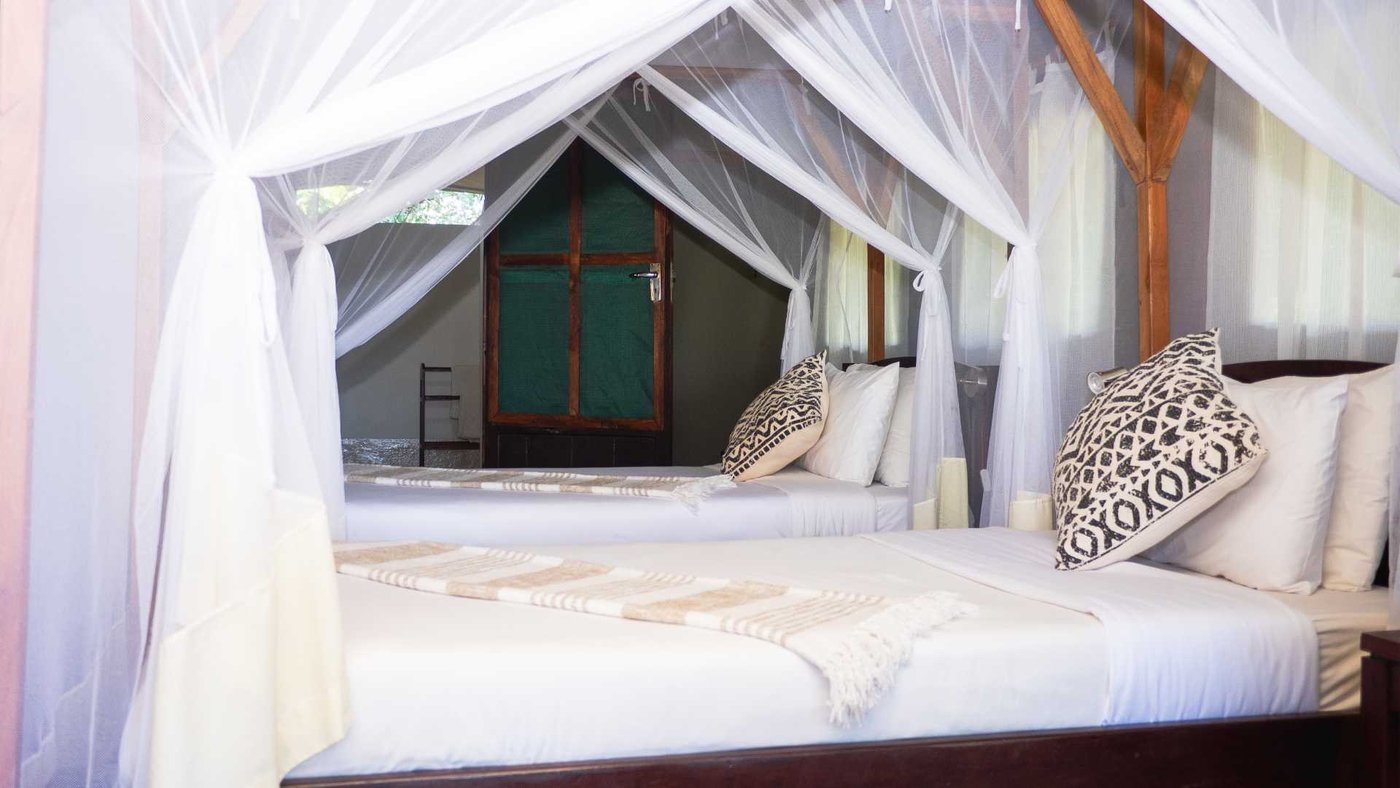 Ein Himmelbett steht unter einem strohgedeckten Pfostendach in einer Suite in Kiambi Safari Lodge.