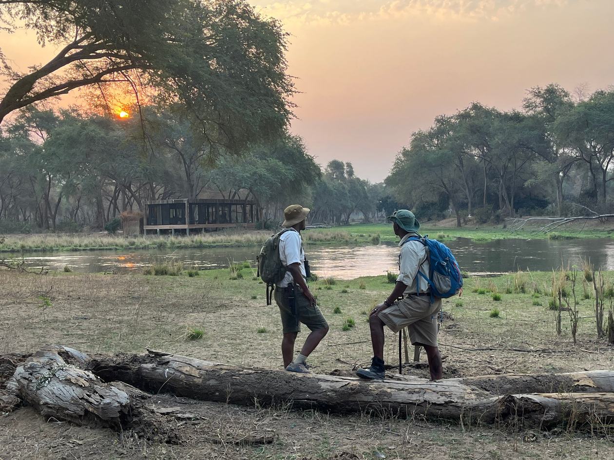 Zwei Guides halten am Fluss bei Sonnenuntergang inne, wobei die Lodge in Lolebezi sichtbar ist.