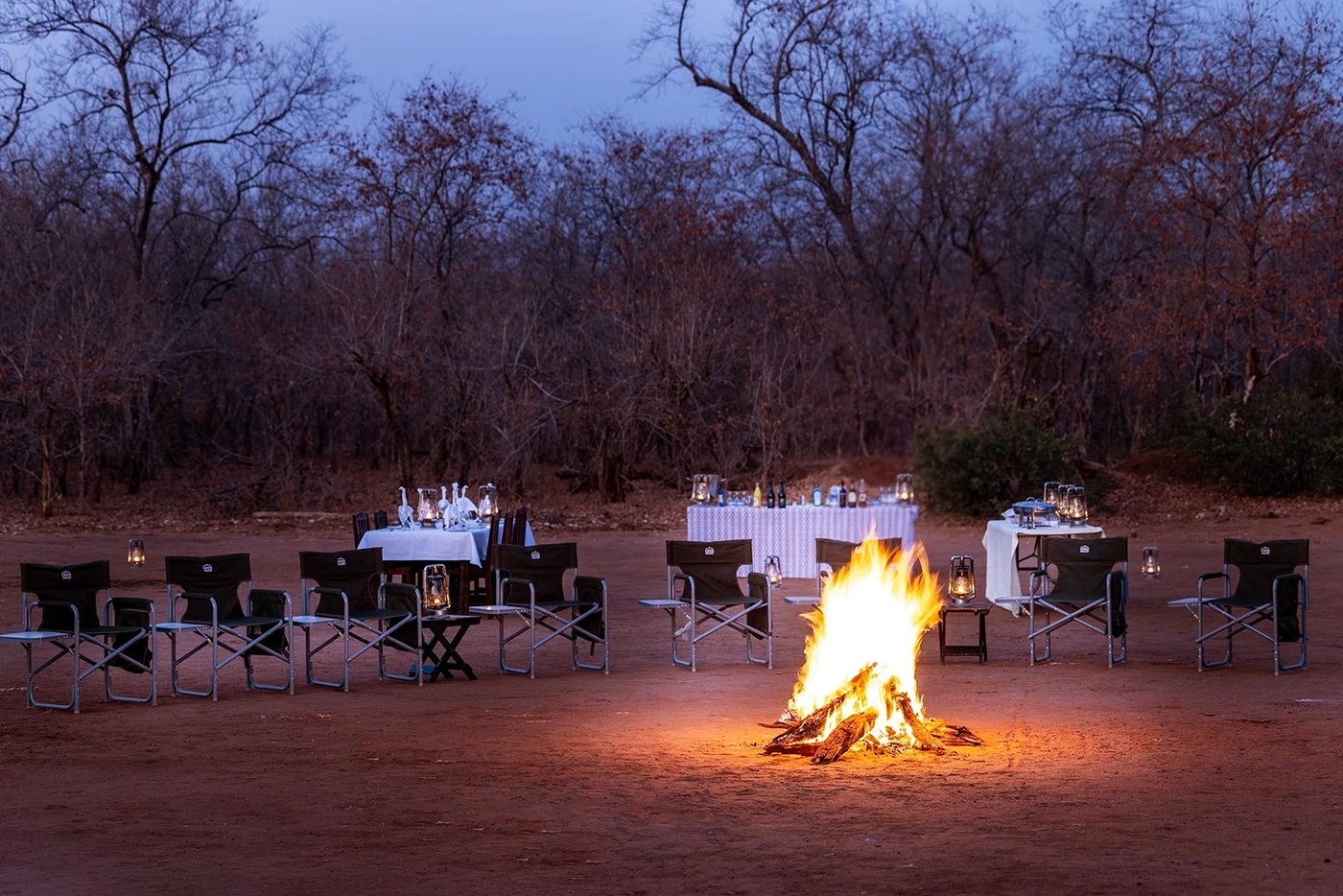 Canvas stoelen en met lantaarns verlichte tafels omcirkelen een boskampvuur voor het diner bij Muchichili Safari House.
