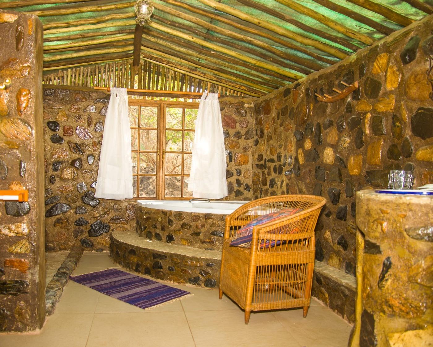 Ein Steinbadezimmer mit eingelassener Badewanne liegt unter einem Strohdach in Mvuu Lodge