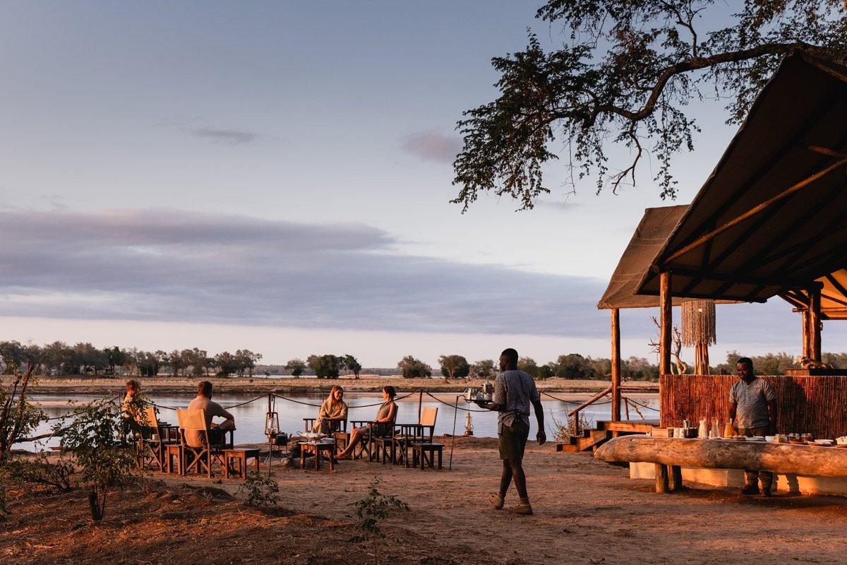 Les clients se rassemblent au boma au bord de la rivière pour l'apéritif pendant qu'un serveur sert les boissons à Old Mondoro.