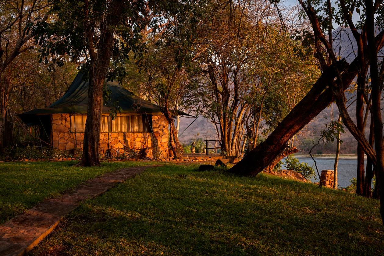 Ein Zeltchalet mit Steinwänden leuchtet bei Sonnenuntergang zwischen Bäumen in der Redcliff Zambezi Lodge in Lower Zambezi